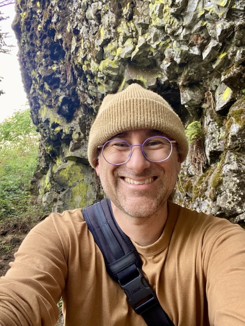 Dott smiling in a tan beanie and lavender glasses, in front of a mossy rock wall.